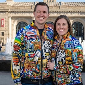 Puede incluir: Dos personas con chaquetas con cremallera a juego con dise&ntilde;os coloridos. Las chaquetas presentan im&aacute;genes de lugares emblem&aacute;ticos, logotipos y el texto "Kansas City 2026". Las chaquetas tienen cuello y pu&ntilde;os negros.