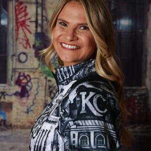 May include: A black and white zip-up jacket with a cityscape design, featuring the letters "KC" prominently displayed. The jacket has a high collar and is worn by a person with blonde hair.