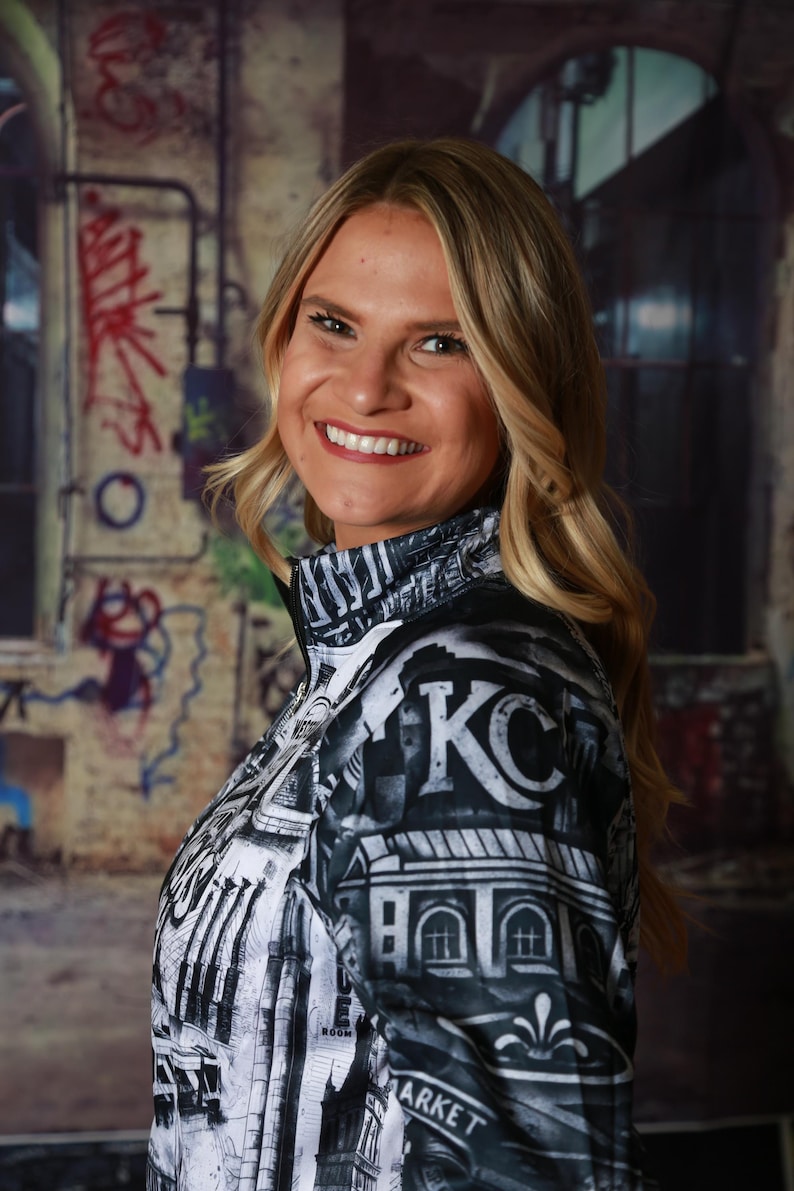 May include: A black and white zip-up jacket with a cityscape design, featuring the letters "KC" prominently displayed. The jacket has a high collar and is worn by a person with blonde hair.