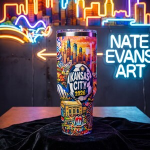 Może przedstawiać: Kubek termiczny ze stali nierdzewnej z kolorowym wzorem panoramy Kansas City, piłką nożną i napisem "Kansas City 2026". Kubek ma srebrną pokrywkę i jest ozdobiony r&oacute;żnymi zabytkami miasta.