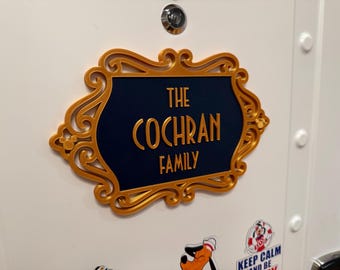 May include: A decorative rectangular sign with a dark blue center and a gold ornate frame featuring small circular accents. The text in gold lettering reads THE COCHRAN FAMILY. The sign is mounted on a white surface below a silver peephole.