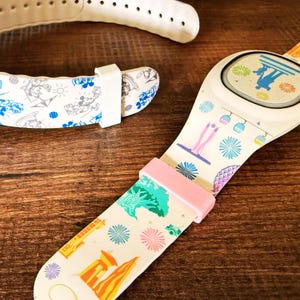 May include: A cream-colored MagicBand wristband with colorful graphics of a theme park. The band features a pink clasp and a variety of illustrations, including fireworks, a tree, and a gondola. Another band and strap are visible in the background.