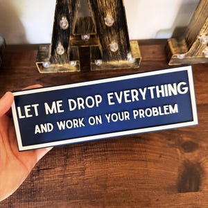 May include: A rectangular navy blue sign with a white border and raised white lettering that reads, LET ME DROP EVERYTHING AND WORK ON YOUR PROBLEM. The sign features a clean, bold font and a flat, matte finish on the surface of the text and frame.