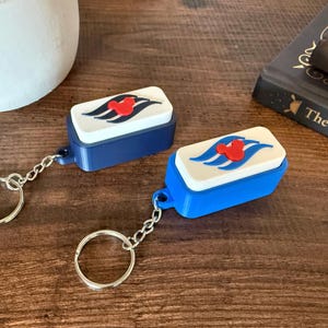 May include: Two rectangular keychains featuring a white top surface with a red Mickey Mouse icon centered above three blue wavy lines. One keychain has a dark blue base and the other has a bright blue base, each with a silver metal split ring.
