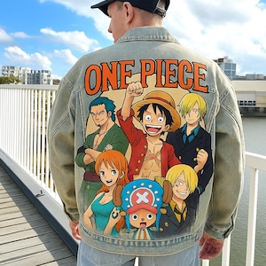 Può includere: Giacca di jeans azzurra con una grafica colorata dei personaggi della serie anime "One Piece". La giacca ha la scritta "ONE PIECE" in arancione sul retro. La giacca è indossata da una persona.
