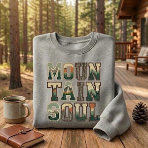 May include: A gray sweatshirt with the words "MOUNTAIN SOUL" in a decorative, nature-themed font. The sweatshirt is folded on a wooden table, next to a coffee mug, a leather-bound journal, and a pine cone. The background features a forest scene.
