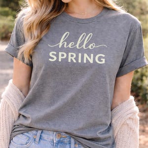 May include: Gray t-shirt with rolled sleeves featuring the words "hello SPRING" in a light yellow script. The shirt is paired with light blue jeans. A cream-colored cardigan is draped over the shoulders.