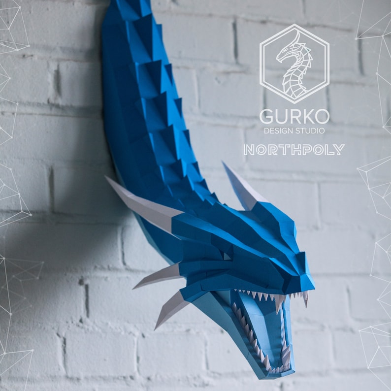 May include: A blue papercraft dragon head with white teeth and horns, mounted on a white brick wall. The dragon's mouth is open and its eyes are black. The text "GURKO DESIGN STUDIO NORTHPOLY" is visible in the background.