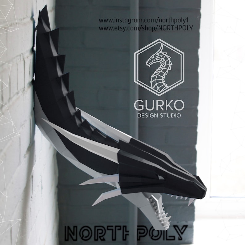 May include: A black and white papercraft dragon head sculpture mounted on a white brick wall. The dragon has a fierce expression with sharp teeth and claws. The sculpture is a 3D model and is part of a papercraft kit. The text "NORTH POLY" is visible at the bottom of the image.