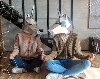 May include: Two people wearing silver unicorn masks are sitting cross-legged on a wooden floor. They are both wearing sweaters and jeans. The person on the left is wearing a light brown sweater and the person on the right is wearing a brown sweater.