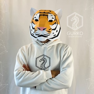 Può includere: Una persona indossa una felpa con cappuccio bianca con un logo di drago nero sul davanti. La persona indossa una maschera di tigre in cartapesta.
