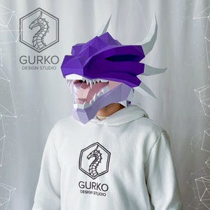 May include: A person wearing a purple and white papercraft dragon mask. The mask has a large open mouth with sharp teeth. The person is wearing a white hoodie with a black dragon logo that says "GURKO DESIGN STUDIO".