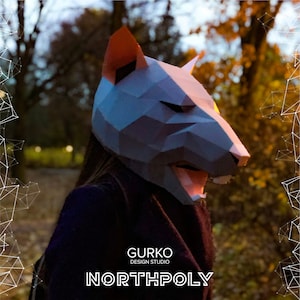 May include: A person wearing a white and grey papercraft mask of a wolf's head. The mask has a sharp, angular design and is partially open, revealing the person's mouth. The background is a blurred image of trees and foliage.