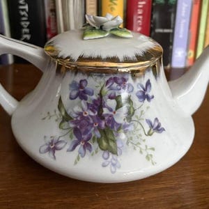 May include: A white porcelain teapot featuring a hand-painted cluster of purple violet flowers with green leaves on the front. The lid is topped with a sculpted white flower and green leaves, and the rim of the lid and spout are accented with gold.