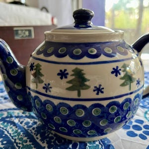 May include: A blue and white teapot with a winter scene design. The teapot has a round body with a blue and white snowflake pattern and green pine trees. The teapot has a blue handle and lid.