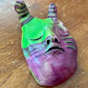 May include: A hand-painted ceramic mask with green and purple paint. The mask has a human-like face with closed eyes and a small nose. The mask has two striped horns on top.