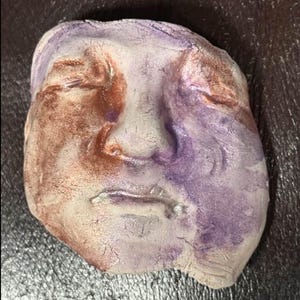 May include: A ceramic sculpture of a face with closed eyes and a downturned mouth. The face is painted in shades of purple, brown, and white.