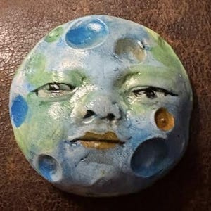 May include: A round, hand-painted sculpture of a face resembling the moon. The face is painted in shades of blue, green, and yellow, with dark lines defining the eyes and mouth. The surface has a textured appearance, with raised areas and indentations.