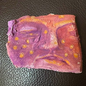 May include: A purple and orange clay sculpture of a face with a sad expression. The face has orange dots on it.