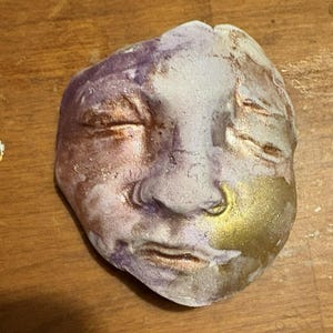 May include: A ceramic face mask with a textured surface. The mask is painted in shades of purple, white, and gold.