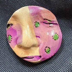 May include: A hand-painted, ceramic face sculpture with a whimsical design. The face is divided into sections of beige, pink, and purple, with green polka dots. Black lines define the eyes and mouth, creating a unique, artistic piece.