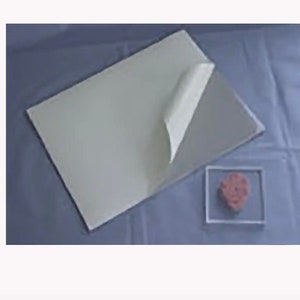 May include: A stack of off-white paper with a corner peeled back, revealing a grey surface underneath. A small, clear square container holds a pink, textured object. The background is a light blue fabric.
