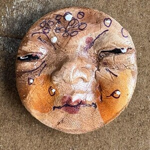 May include: A handmade, round ceramic face with closed eyes, painted with brown, orange, and red hues. The face features black details for the eyes, eyebrows, and mouth, with white accents and a whimsical design on the forehead.