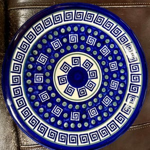 May include: A round, ceramic plate with a vibrant blue and white geometric design. The plate features a repeating Greek key pattern around the rim and a central spiral design. Small green dots are interspersed throughout the blue background.