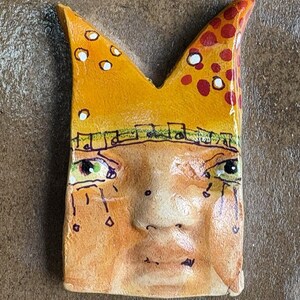 May include: A handmade ceramic art piece featuring a stylized face with a crown. The face is painted in shades of orange and tan, with green and black eyes, and teardrops. The crown is yellow with white and red accents, and a musical note border.