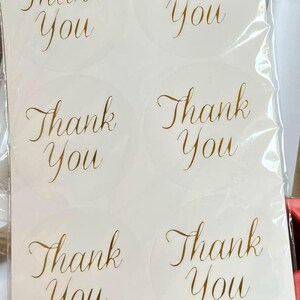 May include: A sheet of white, round stickers with the words "Thank You" in elegant gold script. The stickers are arranged in a grid pattern and are visible through a clear plastic sheet.