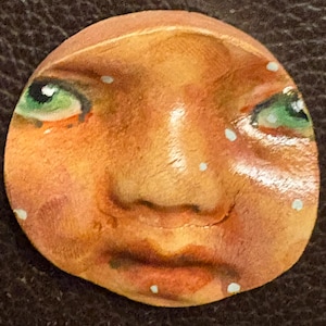 May include: A circular wooden disc featuring a hand-painted face with warm tan skin tones, prominent green eyes, and a neutral expression. The surface is decorated with small, scattered white dots across the cheeks, forehead, and chin area.