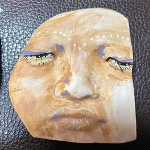 May include: A clay sculpture of a face with green eyes, a purple mouth, and white markings on the forehead.