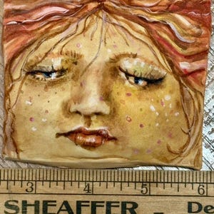 May include: A square ceramic tile featuring a painted face with closed eyes, freckles, and reddish-brown hair. The tile is approximately 2.5 inches wide. A wooden ruler with the text "SHEAFFER HILL, PA." is at the bottom.