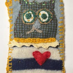 May include: Handmade owl-shaped textile art piece. The owl features green and white circular eyes, a gray beak, and a red heart. The owl is made of various fabrics, including a striped bottom, and is outlined with yellow stitching.