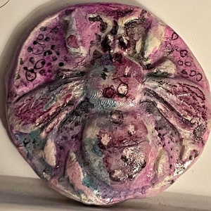 May include: A round, purple, blue, and white ceramic ornament with a bee design. The bee is painted in shades of purple, blue, and white, and has a textured surface. The ornament is decorated with black dots and lines.