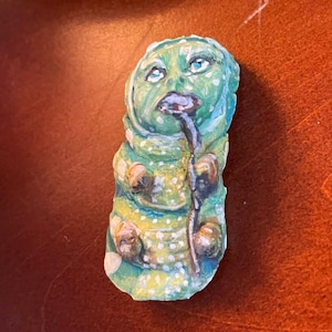 May include: A green and white hand-painted ceramic figurine of a caterpillar with a large head and a long, thin body. The caterpillar has a large, round head with a wide mouth and large, blue eyes. The caterpillar is wearing a green and white patterned hat.