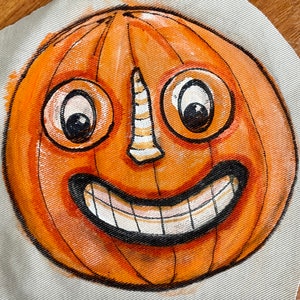May include: A hand-painted orange pumpkin with a wide, toothy grin, large eyes, and a white nose. The pumpkin is painted on a white canvas.