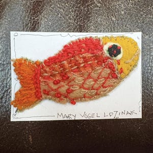 May include: Handmade felt fish art piece with red, orange, and yellow hues. The fish has a detailed, textured design with stitched accents and a black and red eye. The artist's name, Mary Vogel Lozinak, is written below the fish.