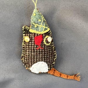 May include: A brown and white felt cat ornament with a green and yellow hat. The cat has a red nose and white eyes with yellow accents.