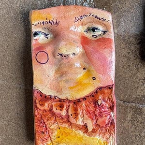 May include: A rectangular, handmade art piece featuring a stylized face with a whimsical design. The face has a peach complexion with red cheeks, black markings, and a red-orange beard. A metal loop is attached at the top for hanging.