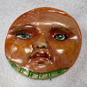 May include: A circular, glossy ceramic wall plaque featuring a sculpted human face with prominent cheeks, a small nose, and detailed green eyes. The skin tone is a warm, mottled brown with orange highlights, and a green textured band lines the bottom.