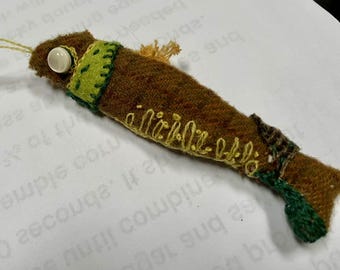 May include: A brown felt fish with green and yellow embroidery. The fish has a white button eye and a small yellow felt fin.