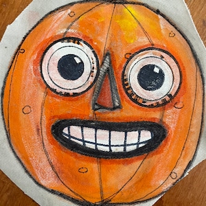 May include: A hand-painted orange pumpkin face with large eyes, a triangular nose, and a wide grin. The artwork is on fabric, possibly for a Halloween decoration or craft project. The style is whimsical and folk-art inspired, perfect for autumn.