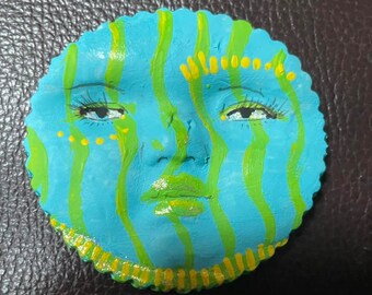 May include: A round, blue and green painted ceramic ornament with a face. The face has yellow painted eyes, nose, and mouth.