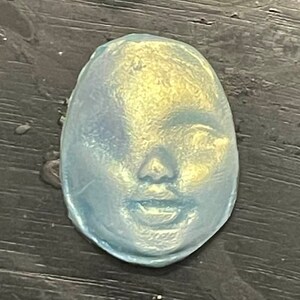 May include: A blue and gold oval-shaped resin sculpture of a face with a neutral expression.