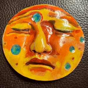 May include: A circular wall plaque featuring a sculpted human face with closed eyes, a prominent nose, and full lips. The surface is painted in vibrant shades of orange and yellow, accented with small, scattered blue and green circular dots.