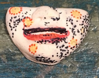 May include: A white ceramic face with a black and orange painted design. The face has a closed mouth and black eyes. The design includes black dots and orange circles.