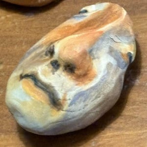 May include: A ceramic sculpture of a face with a textured surface. The face is painted in shades of brown, orange, and gray. The sculpture has a distressed look, as if it has been weathered by time.