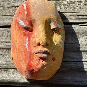 May include: A ceramic mask with a textured surface, painted in shades of orange, yellow, and white. The mask has a stylized human face with closed eyes and a slightly open mouth.