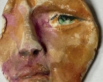May include: A ceramic sculpture of a face with a textured surface. The face is painted in shades of brown, pink, and green. The sculpture has a distressed look, with cracks and chips in the surface.